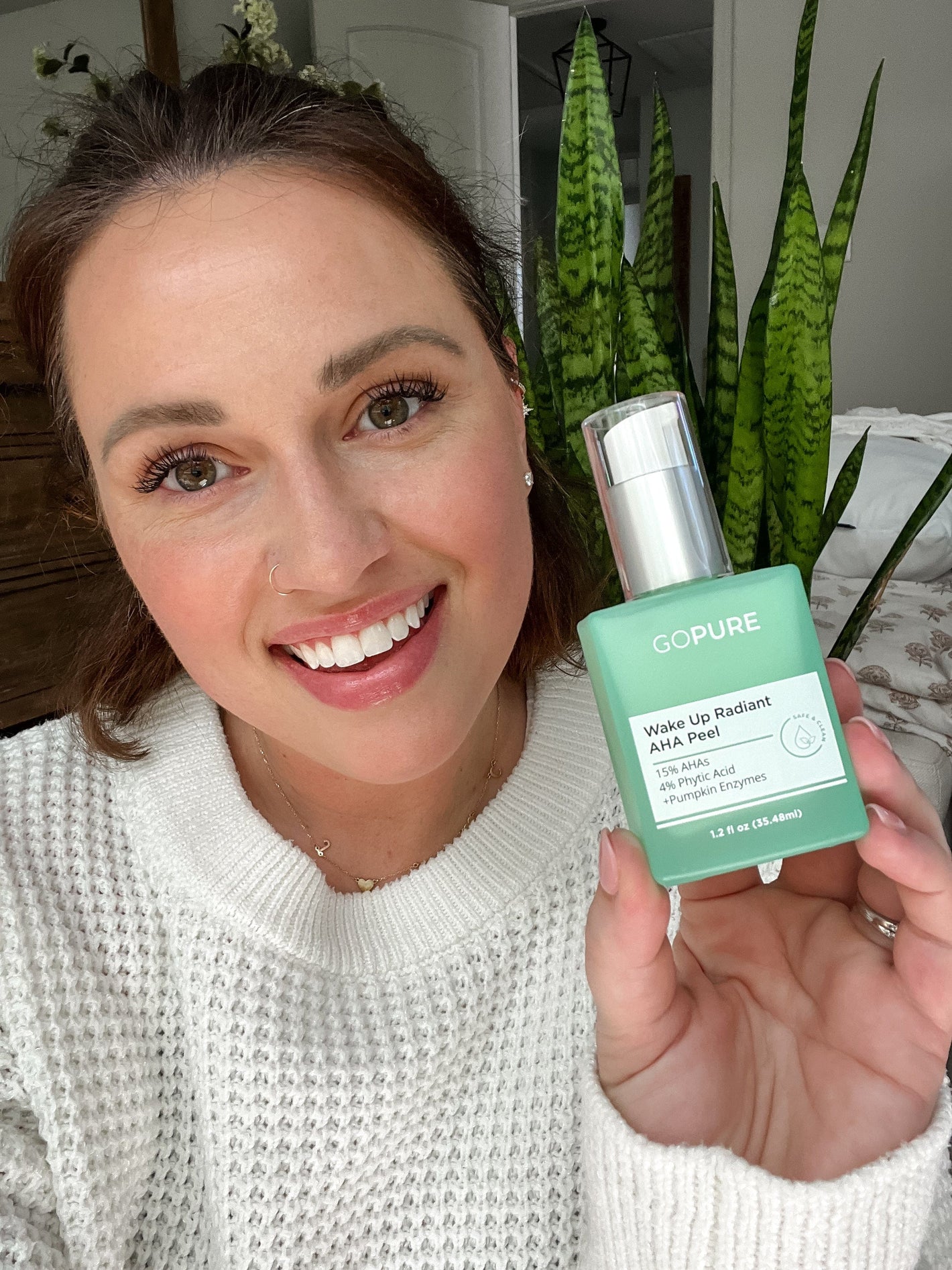 A smiling woman holding a green bottle of "GoPure Wake Up Radiant AHA Peel" in her hand, with text highlighting 15% AHA, 5% PHA, and Pumpkin Enzyme. She is indoors with a vibrant snake plant in the background, emphasizing the skincare product.