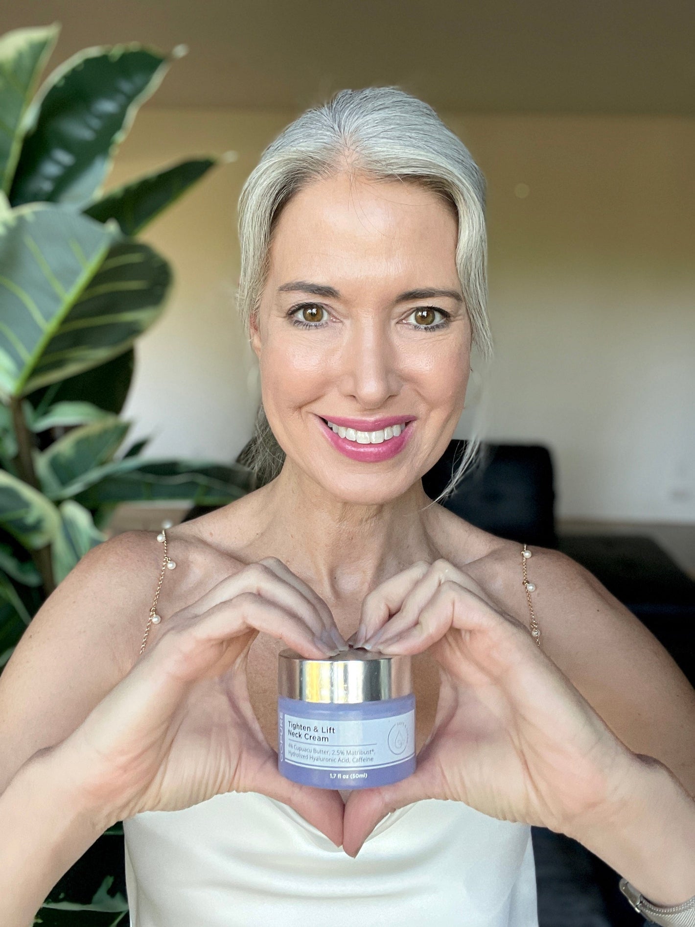 A smiling woman holding a jar of skincare cream in her hands, with a lush green plant in the background. She appears in a well-lit indoor space, emphasizing the product she’s showcasing.