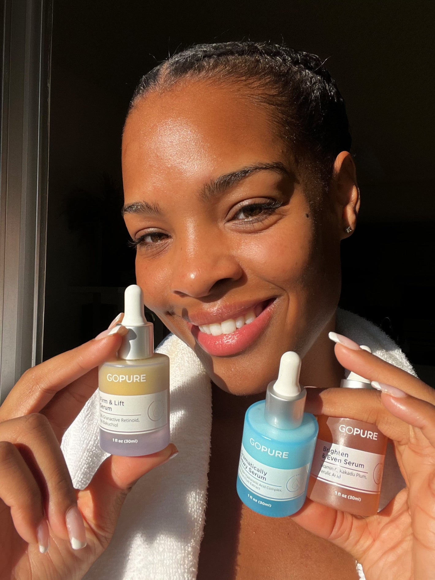 A smiling woman with smooth skin holds three bottles of GoPure skincare serums close to her face. She is wearing a white robe, and the bottles are labeled "Firm & Lift Serum," "Hyaluronic Acid Serum," and "Brighten & Even Serum.