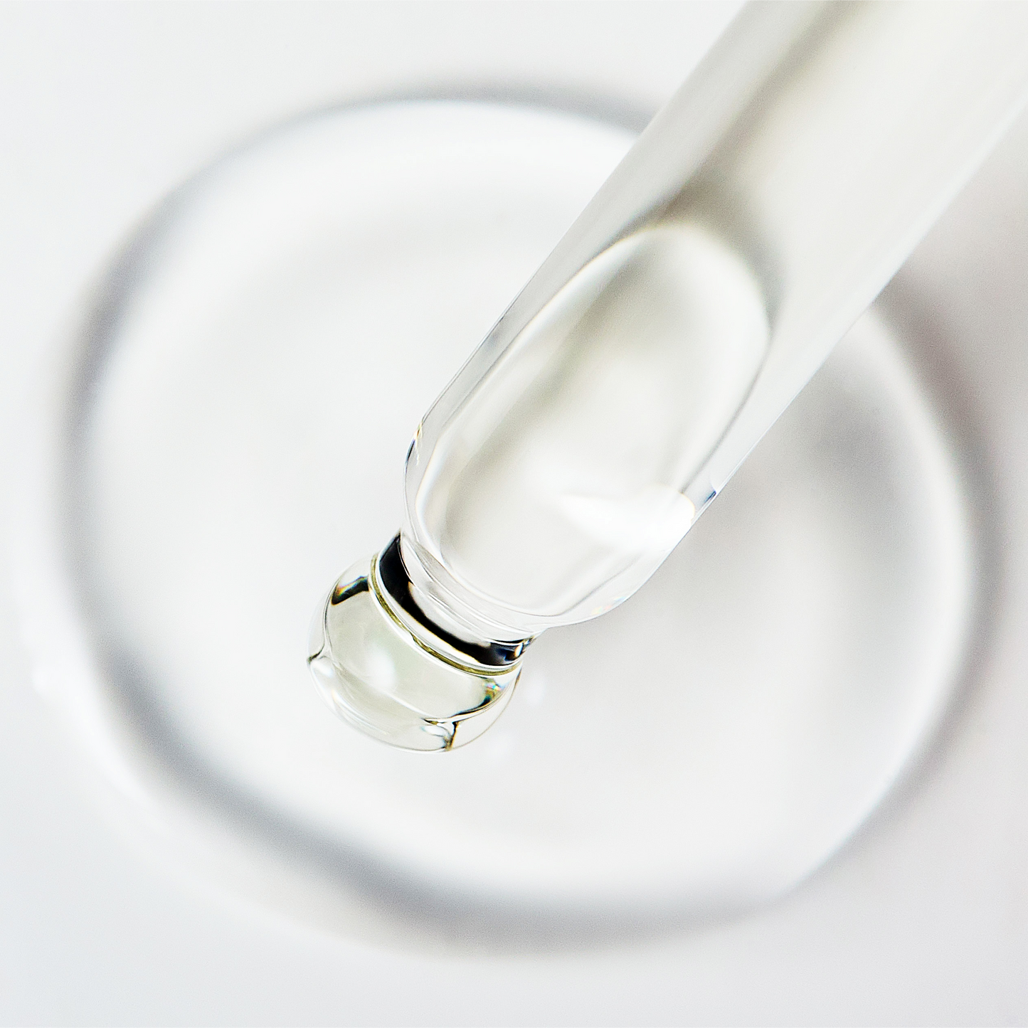 Close-up image of a clear serum dropper releasing a droplet of transparent liquid onto a smooth white surface showing the glossy texture of the skincare formula