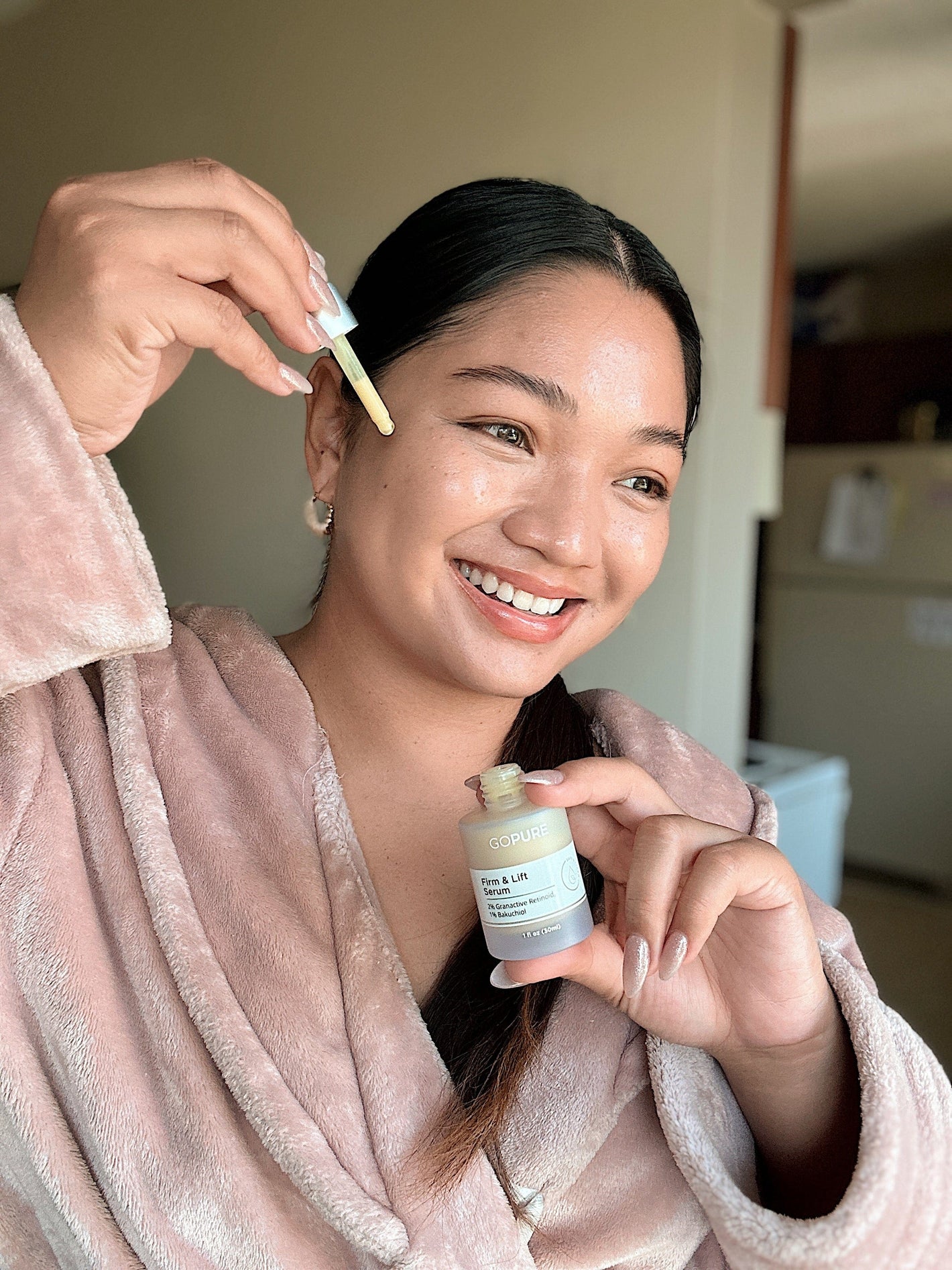 Smiling woman applying GoPure Firm & Lift Serum to her face using a dropper. She is wearing a light pink robe and is posed in a cozy indoor setting.