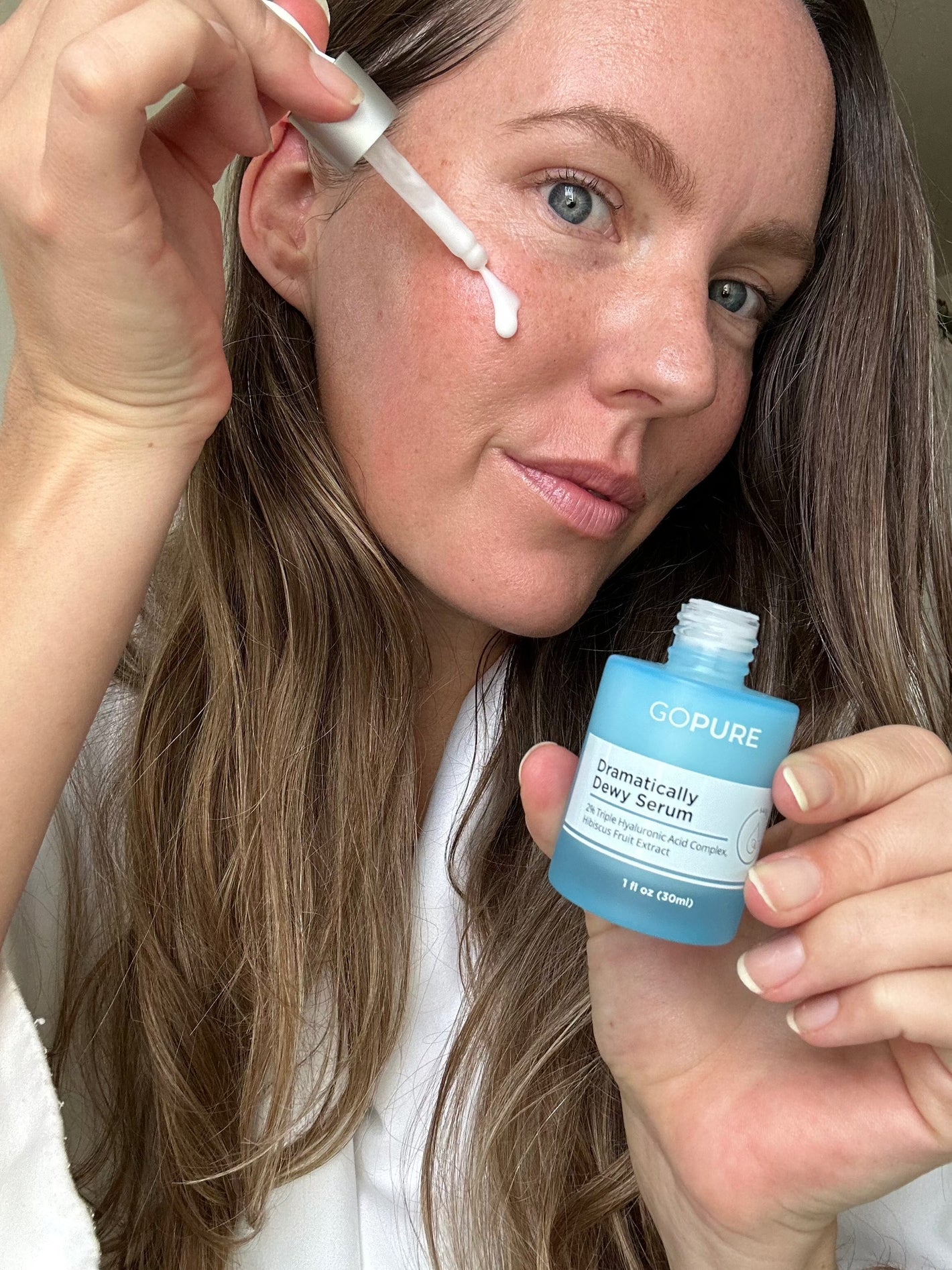 A woman with long brown hair applies GoPure's Dramatically Dewy Serum to her cheek using a dropper. She holds the blue serum bottle in her other hand, looking at the camera with clear skin and a slight smile.