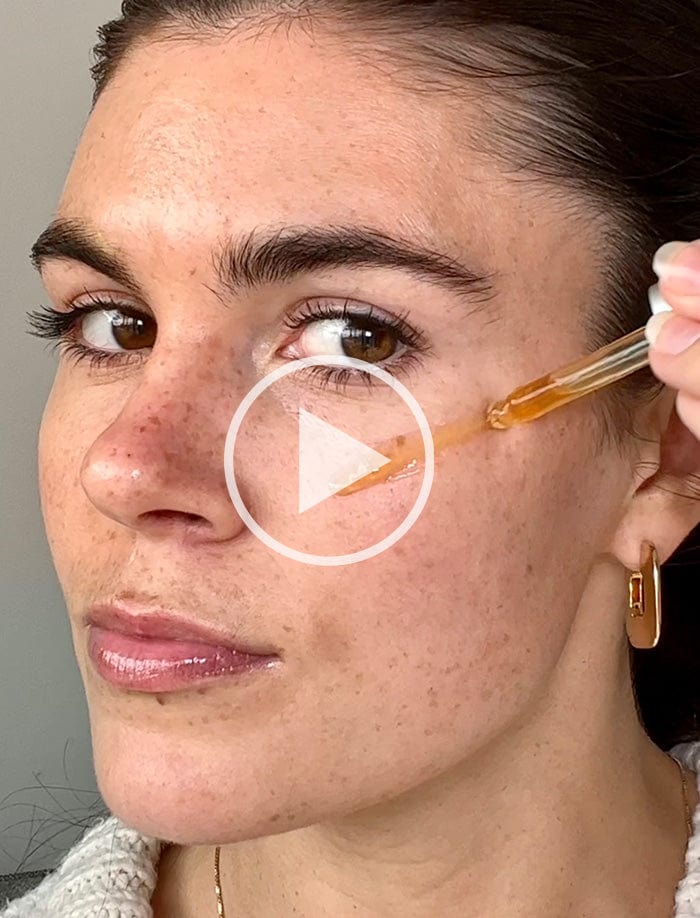 Woman applying serum to her face using a dropper, with focus on her cheek. She has brown eyes, dark hair, and is wearing gold earrings. A play button icon is overlaid on the image, indicating a video.