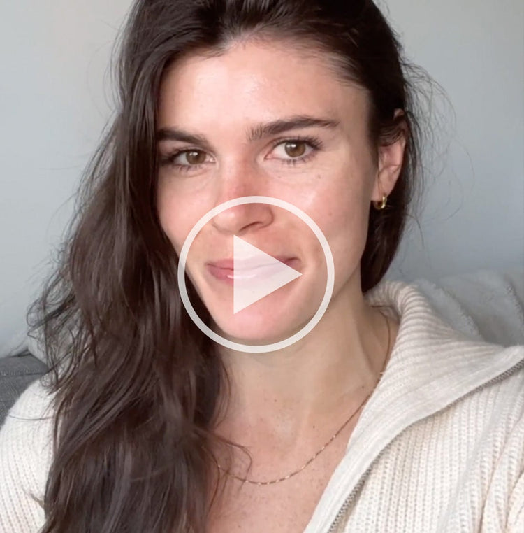 Woman with long dark hair and light skin wearing a white sweater, smiling and looking at the camera with a play button overlay, indicating a video.