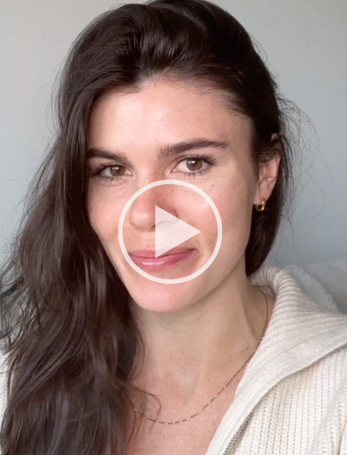 Woman with long dark hair and light skin wearing a white sweater, smiling and looking at the camera with a play button overlay, indicating a video.