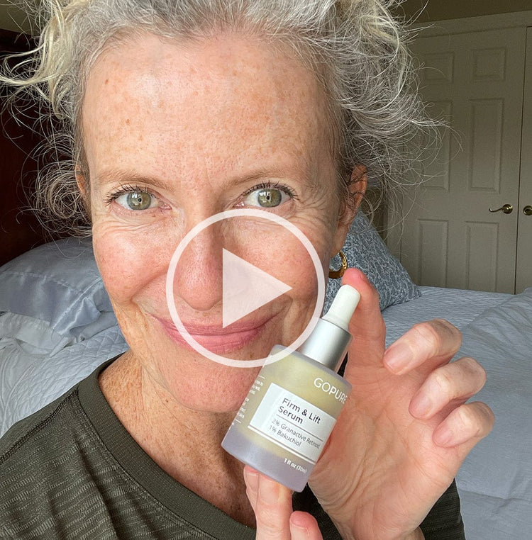 Smiling woman with gray hair holding GoPure Firm & Lift Serum. A play button icon is overlaid, indicating a video. She is in a bedroom setting.