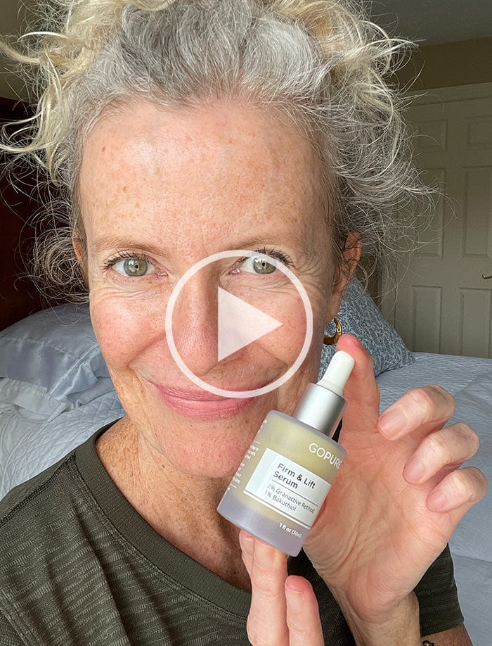 Smiling woman with gray hair holding GoPure Firm & Lift Serum. A play button icon is overlaid, indicating a video. She is in a bedroom setting.
