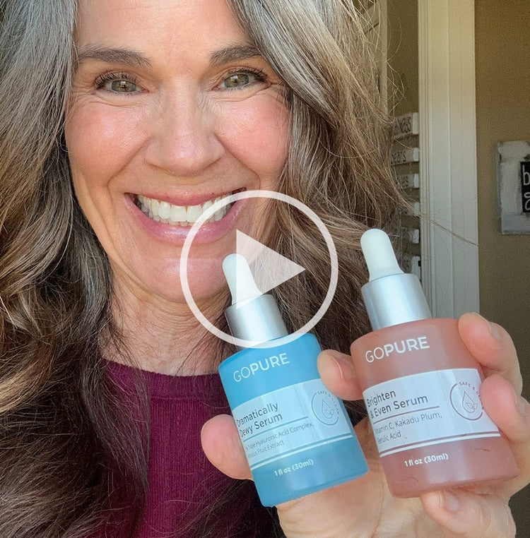 Smiling woman with gray hair holding GoPure Dramatically Dewy Serum and Brighten & Even Serum. A play button icon is overlaid, indicating a video.