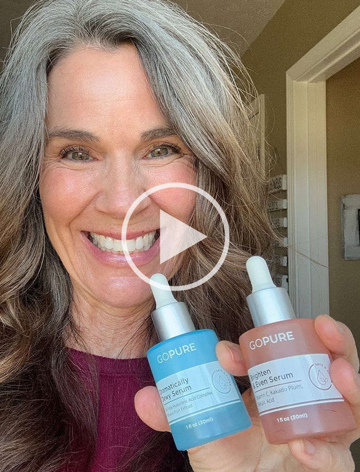 Smiling woman with gray hair holding GoPure Dramatically Dewy Serum and Brighten & Even Serum. A play button icon is overlaid, indicating a video.