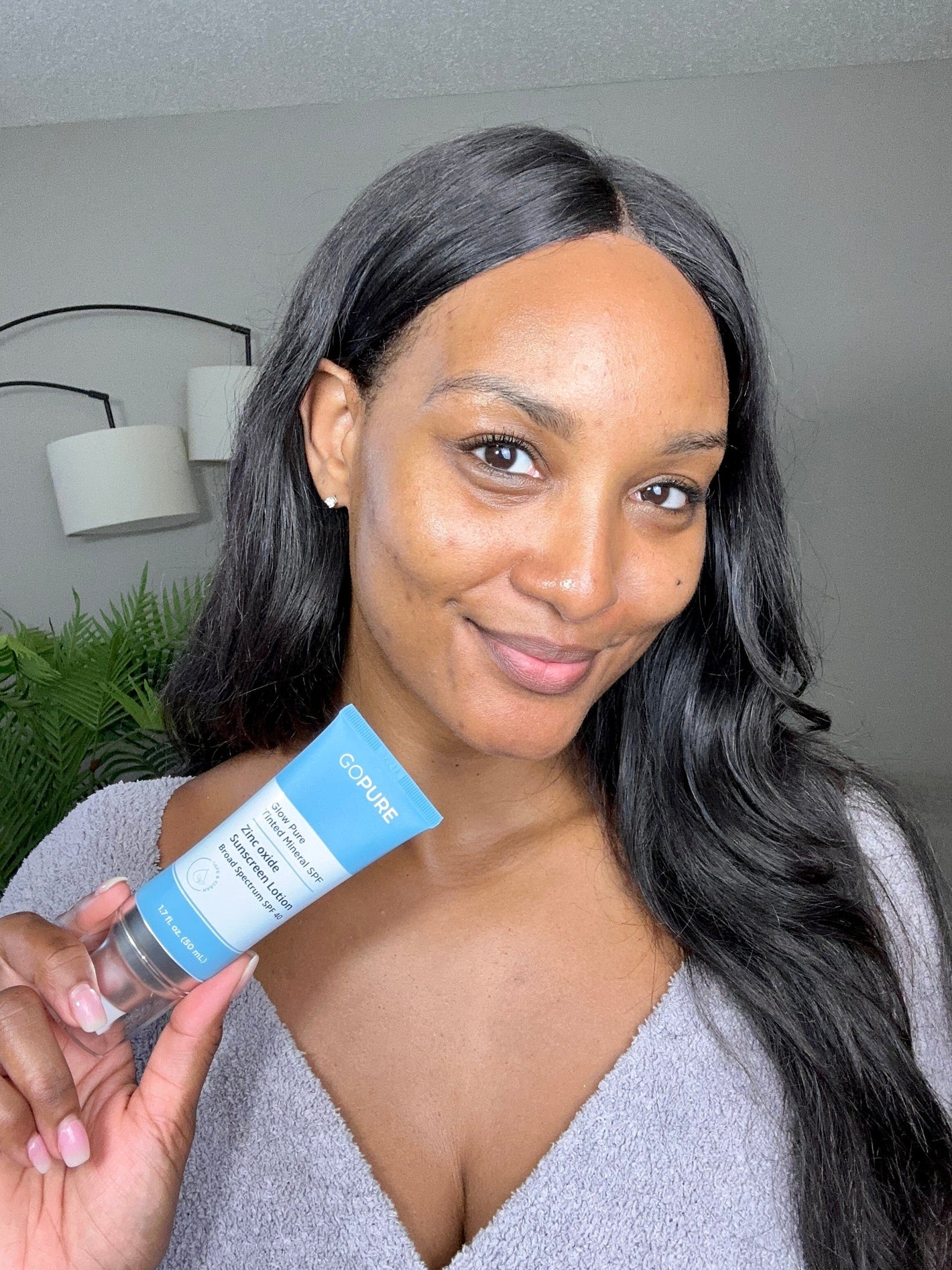  A woman with long black hair holds a tube of GoPure Actives Intensive Spot Corrector, smiling softly. She has clear skin and is wearing a light gray sweater. Indoor setting with green plants in the background.