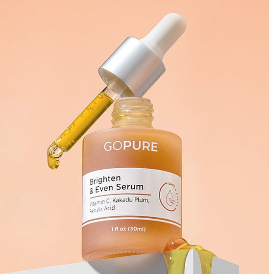 Bottle of GoPure Brighten & Even Serum with a dropper leaning on the bottle, dispensing yellow serum. The label indicates it contains Vitamin C, Kakadu Plum, and Ferulic Acid. The background is a gradient of light peach.