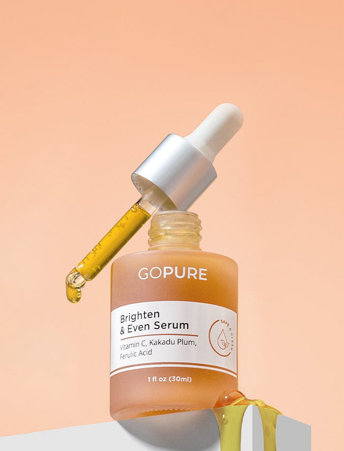 Bottle of GoPure Brighten & Even Serum with a dropper leaning on the bottle, dispensing yellow serum. The label indicates it contains Vitamin C, Kakadu Plum, and Ferulic Acid. The background is a gradient of light peach.