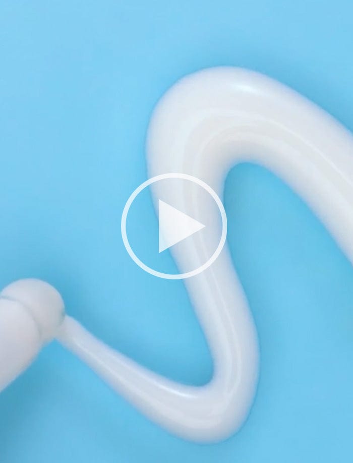 White cream being dispensed in a wavy line on a blue surface. A play button icon is overlaid on the image, indicating a video.