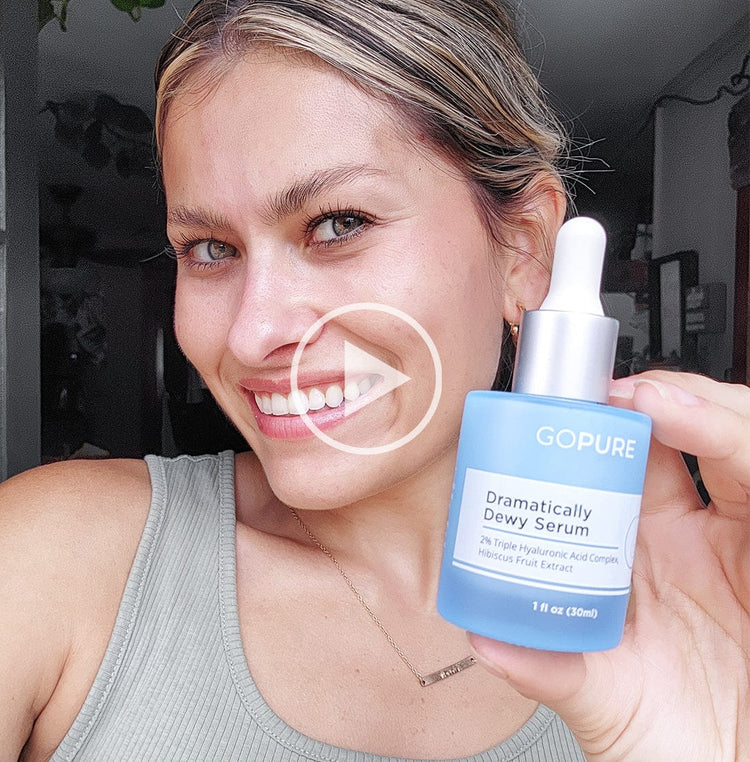 Smiling woman holding a bottle of GoPure Dramatically Dewy Serum. She is wearing a gray tank top and posed indoors. A play button icon is overlaid on the image, indicating a video.