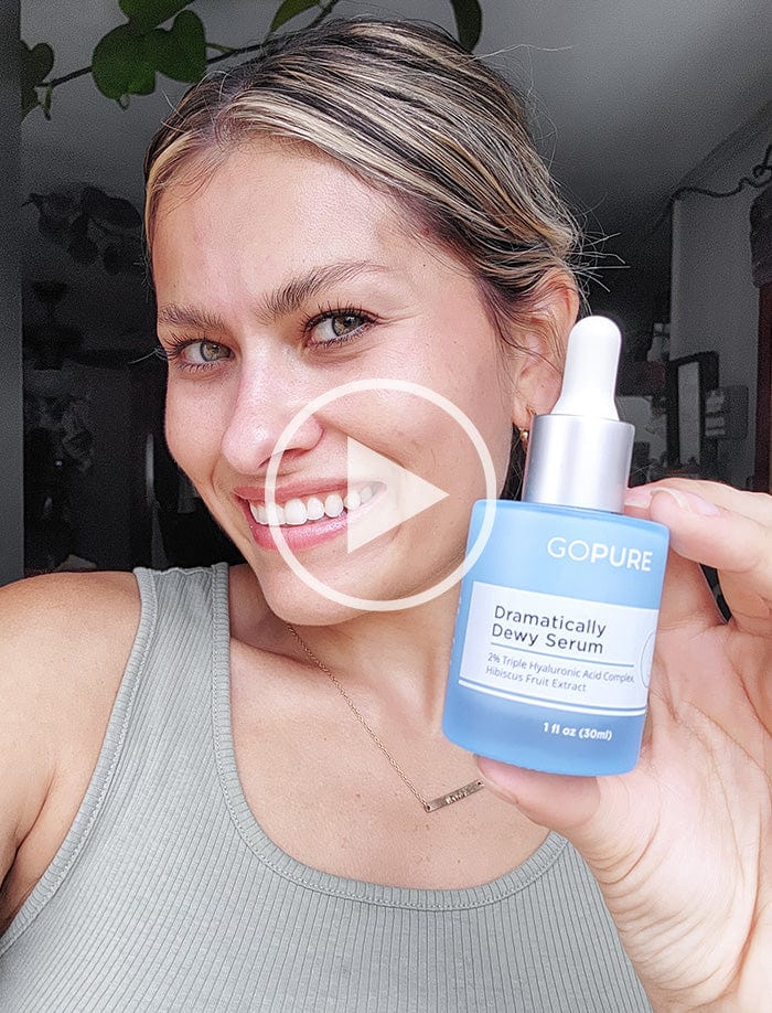 Smiling woman holding a bottle of GoPure Dramatically Dewy Serum. She is wearing a gray tank top and posed indoors. A play button icon is overlaid on the image, indicating a video.