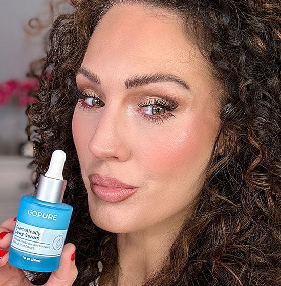 Woman with curly hair holding a bottle of GoPure Dramatically Dewy Serum. She has a slightly pouty expression and is posed in a well-lit indoor setting.