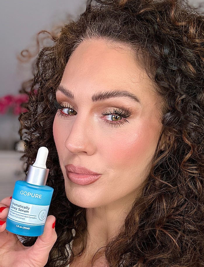 Woman with curly hair holding a bottle of GoPure Dramatically Dewy Serum. She has a slightly pouty expression and is posed in a well-lit indoor setting.