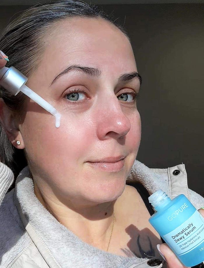 Woman applying GoPure Dramatically Dewy Serum to her face using a dropper. She is holding the serum bottle and wearing a cozy fleece jacket, posed in a well-lit indoor setting.