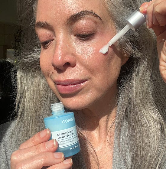 Woman with long gray hair applying GoPure Dramatically Dewy Serum to her face using a dropper. She is holding the serum bottle and smiling slightly, posed in a well-lit indoor setting.