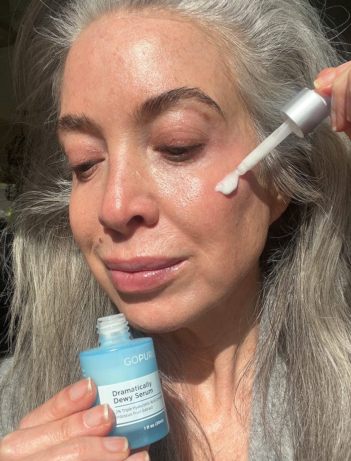 Woman with long gray hair applying GoPure Dramatically Dewy Serum to her face using a dropper. She is holding the serum bottle and smiling slightly, posed in a well-lit indoor setting.