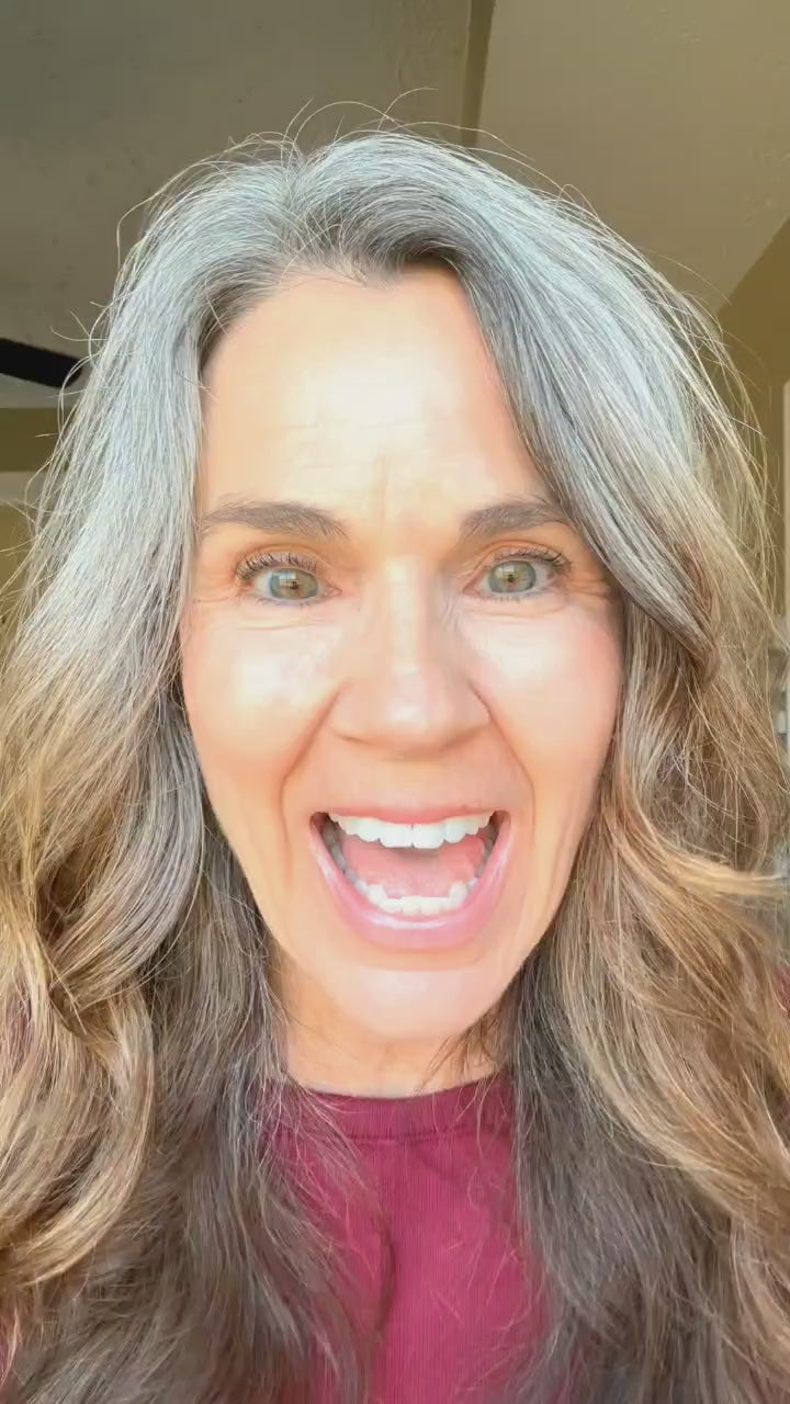Woman with long gray hair and bright eyes smiling widely, appearing excited in a well-lit indoor setting.