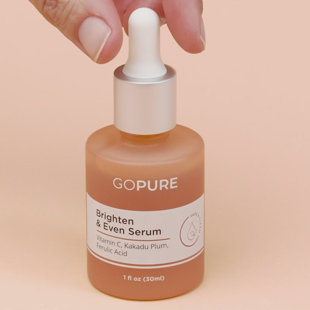 Hand holding the dropper of a GoPure Brighten & Even Serum bottle with Vitamin C, Kakadu Plum, and Ferulic Acid, on a peach-colored background.